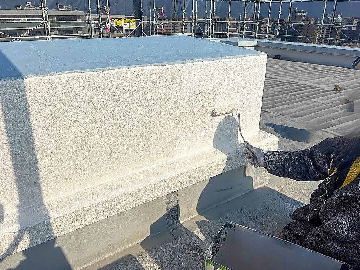 屋上防水層へのスプレー塗装施工。作業者が大型の白い防水材料に対して遠隔スプレーガンで塗装を行っており、都市部の高層建築の屋上環境が背景。